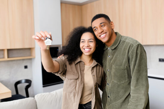 Happy young married couple holding a key ring in hand, hugging and looking at it with smile, while standing in the modern kitchen of their new home. Excited owners of a condo